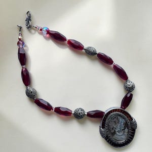 Pode incluir: Um colar de contas bordô com um pingente de camafeu. O colar apresenta contas bordô facetadas, contas prateadas ornamentadas e um pingente de camafeu com um retrato de perfil. O colar é preso com um fecho prateado.