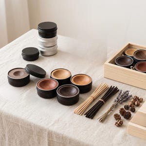 Puede incluir: Un surtido de pequeños recipientes redondos con tapas negras e inserciones de madera. Algunos recipientes están apilados, otros están abiertos, revelando varios tonos de madera. También se disponen manojos de varillas de incienso, lavanda seca y piñas.