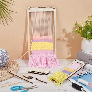 Peut inclure: Un métier à tisser en bois avec des fils de chaîne blancs verticaux et un textile partiellement tissé présentant des bandes horizontales de laine violette, jaune et rose avec une longue frange rose, accompagné d'outils de tissage.