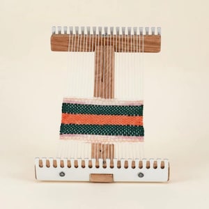 May include: A wooden weaving loom featuring a vertical frame with white plastic notched bars at the top and bottom. White warp threads are strung vertically, holding a partially woven textile with horizontal stripes in pink, dark green, and orange.