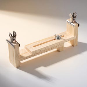 May include: A light-coloured wooden adjustable jig featuring two silver metal binder clips attached to vertical end posts. The base includes a horizontal slot with a metal wing nut and a printed ruler scale ranging from 15 to 30 centimetres.