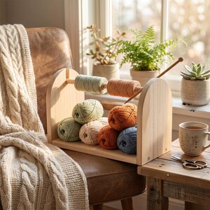 May include: A wooden yarn organizer holds various colors of yarn and thread. The yarn is in shades of green, orange, and blue. A wooden knitting needle is inserted into a spool of orange thread. A cream-colored cable-knit blanket is draped over a chair.