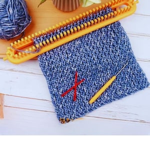 Puede incluir: Un telar de punto amarillo con hilo azul y blanco en proceso. Una bola de hilo azul y blanco está a la izquierda. Un ganchillo amarillo y una cruz roja están en la tela tejida. El fondo es una superficie de madera clara.