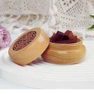 Op de afbeelding: Een lichtbruine houten wierookdoos met een decoratief deksel, open om kleine, donkere houten blokken te laten zien. De doos heeft een gesneden ontwerp op het deksel. De doos staat op een witte tray, wat wijst op aromatherapie of huisgeur.