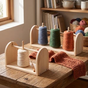 May include: Wooden yarn swift with a white yarn cone, a crochet hook, and a rust-colored knitted scarf. Other swifts hold blue, green, and orange yarn cones. A basket of yarn balls sits on a shelf in the background.