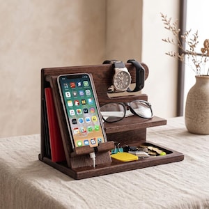 Op de afbeelding: Een donkerbruine houten bureau-organizer met meerdere compartimenten. Het bevat een smartphone, een horloge, een fitnesstracker, een bril, sleutels en munten. Een rood notitieboek is zichtbaar. De organizer staat op een licht oppervlak.