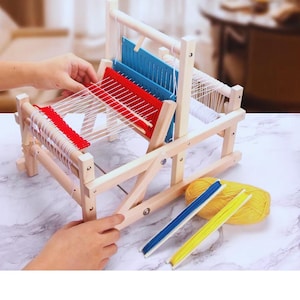 May include: A wooden weaving loom with red and blue thread guides. The loom is partially assembled with white thread and includes a ball of yellow yarn and two threaders. The loom is on a marble surface.