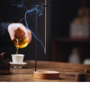 May include: A wooden incense holder with a black metal stand, holding a lit incense stick. Wisps of smoke curl upwards. The holder sits on a wooden surface, with a cup and teapot in the background, suggesting a tea ceremony.