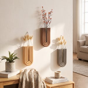 May include: Three wall-mounted, U-shaped vases in varying shades of brown and gray, each holding dried flowers. The vases have a vertical, grooved design and are suspended by metal wires. A small potted plant and a stack of books are visible below.