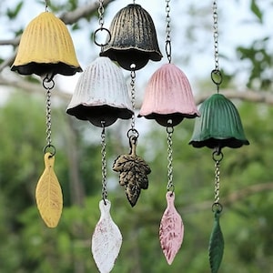 May include: A selection of hanging wind chimes with bell-shaped tops in various colours, including yellow, black, white, pink, and green. Each chime is suspended by a chain and features a leaf-shaped or leaf-design pendant. The background is a blurred view of foliage.