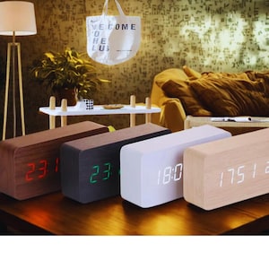 Puede incluir: Una colección de relojes digitales rectangulares de madera en varios tonos. Cada reloj muestra la hora en formato digital, con los números iluminados en rojo, verde y blanco. Los relojes están colocados sobre una superficie de madera oscura.