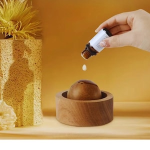 Può includere: Un diffusore di oli essenziali in legno con una parte superiore marrone arrotondata e una base circolare. Una mano versa olio da una piccola bottiglia marrone con un'etichetta bianca. Lo sfondo è di un caldo colore dorato.