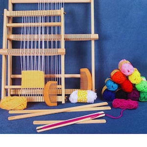 Op de afbeelding: Een houten weefgetouwset met twee weefgetouwen, een kam en diverse garenkleuren. De set bevat geel, oranje, roze, blauw en groen garen. De weefgetouwen zijn gemaakt van lichtgekleurd hout tegen een blauwe achtergrond.