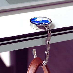 May include: A silver handbag hook with a blue and white floral design. The hook is attached to a brown leather handbag. The hook is designed to hang a handbag from a table or desk.