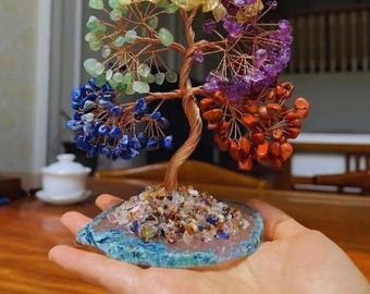May include: A decorative gemstone tree with a copper wire trunk and branches. The tree features colorful gemstones in shades of yellow, green, blue, purple, and orange. The base is a blue-edged stone, and the tree is held in a hand.