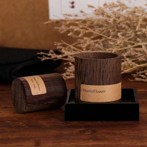 Puede incluir: Un recipiente cilíndrico de madera marrón oscuro para difusor de varillas con una etiqueta beige que dice HomeFlower, colocado sobre una base cuadrada negra. Al lado hay un cilindro de madera a juego con la etiqueta Reed Diffuser Since 2009.