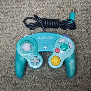 May include: Teal Nintendo GameCube controller with a black cord and teal plug. The controller has a directional pad, analog stick, and various buttons in red, green, and yellow. The text "NINTENDO GAMECUBE" is printed on the controller.