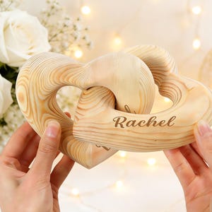 May include: Two interlocking heart-shaped wooden ornaments made of light-coloured pine with visible wood grain. The front heart features the name Rachel engraved in a dark brown script font. The smooth, polished surface highlights the natural texture.