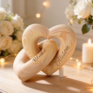 May include: A light brown wooden heart sculpture with interlocking loops, engraved with the names "Blake" and "Alison". The sculpture is on a wooden surface, with white roses and candles in the background.