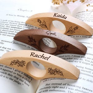 Puede incluir: Tres sujetapáginas de madera con los nombres personalizados "Kaida", "Sophie" y "Rachel". Cada uno presenta un diseño floral y un agujero para el dedo. Los sujetapáginas están sobre las páginas de un libro abierto.