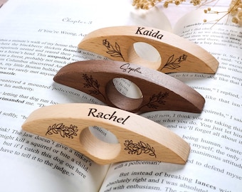 May include: Three wooden thumb page holders with personalized names: "Kaida", "Sophie", and "Rachel". Each holder features a floral design and a finger hole. The holders are resting on the pages of an open book.