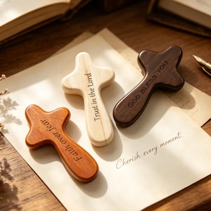 Könnte beinhalten: Drei Holzkreuze in verschiedenen Braun- und Naturholztönen werden auf einem Papier gezeigt. Die Kreuze tragen die eingravierten Texte: "Faith over fear", "Trust in the Lord" und "God bless you". Ein Stift und ein offenes Buch befinden sich im Hintergrund.