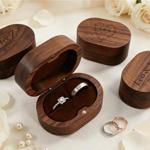 May include: Four oval dark brown wooden ring boxes with hinged lids. Three boxes are closed and engraved with names and dates. One box is open, revealing a dark brown velvet interior holding two silver-toned rings with clear stones.