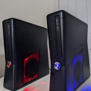 May include: Two black Xbox 360 consoles are shown. One has a red light emitting from its vent, the other a blue light. The Xbox 360 logo is visible on the top of each console. The consoles are on a white surface.