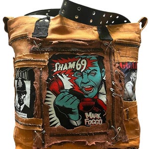 May include: A brown canvas tote bag with multiple patches and a black belt strap. The patches feature graphics and text, including "SHAM 69" and "MARK FOGGO". The bag has a distressed, punk rock aesthetic.