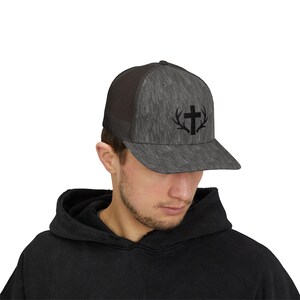 Könnte beinhalten: Graue Trucker-Mütze mit schwarzem Kreuz- und Geweih-Design. Die Mütze hat eine Mesh-Rückseite und einen gebogenen Schirm. Die Vorderseite hat ein Holzmaserungsmuster.
