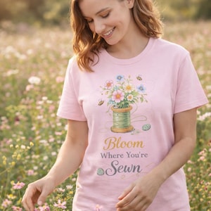 Puede incluir: Camiseta rosa claro con un diseño floral y de abejas, y el texto "Bloom Where You're Sewn". La camiseta presenta una bobina de hilo con flores y abejas. La persona está en un campo de flores.