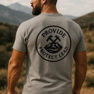 May include: A gray t-shirt with a circular black graphic on the back. The graphic includes crossed pickaxes and the words "PROVIDE", "PROTECT", and "LEAD". The shirt is short-sleeved and worn by a person outdoors.