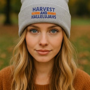 May include: A light gray beanie with the embroidered text "HARVEST AND HALLELUJAHS" in blue and orange. The beanie is worn by a person with blonde hair and blue eyes, wearing a brown sweater. The background is blurred with fall colors.