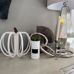 Può includere: Una composizione decorativa con una zucca bianca a telaio aperto con uno stelo marrone, un vaso geometrico bianco con un'etichetta nera e una scultura a catena d'argento. Sono presenti anche una candela accesa e una lampada decorativa.