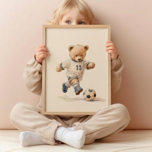May include: A rectangular framed print featuring a watercolor illustration of a light brown teddy bear wearing a white sports jersey with the number 13 in dark blue, white shorts, and blue sneakers, actively kicking a black and white soccer ball.