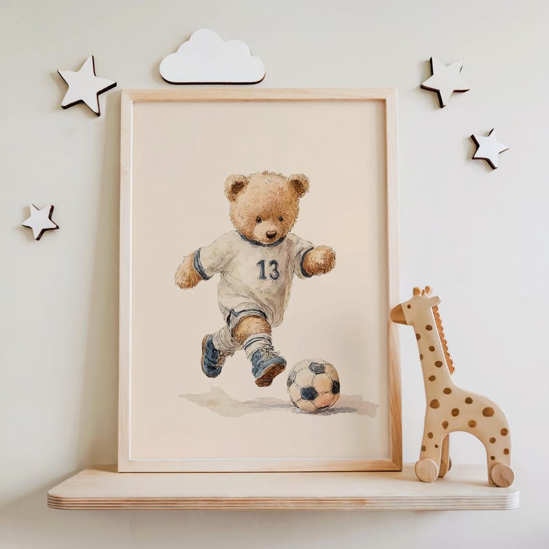 May include: A framed watercolor print featuring a light brown teddy bear wearing a white soccer jersey with the number 13 in blue, blue shorts, and blue soccer cleats, actively kicking a black and white soccer ball on a light beige background.