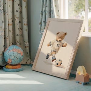 May include: A framed watercolor-style print featuring a light brown teddy bear wearing a white soccer jersey with the number 13 in blue, running and kicking a soccer ball. The artwork is presented in a light-colored wooden frame.