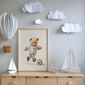 May include: A framed watercolor print of a teddy bear wearing a white soccer jersey with the number 13, kicking a soccer ball. The scene includes a hanging paper hot air balloon, three white paper clouds, and two white model sailboats.