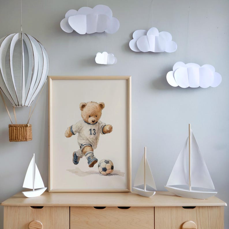 May include: A framed watercolor print of a teddy bear wearing a white soccer jersey with the number 13, kicking a soccer ball. The scene includes a hanging paper hot air balloon, three white paper clouds, and two white model sailboats.