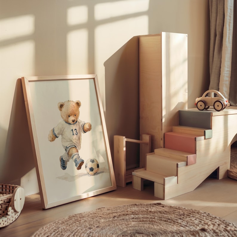 May include: A framed print featuring a light brown teddy bear wearing a white soccer jersey with the number 13 and blue shorts, kicking a soccer ball. Next to it is a light-colored wooden staircase structure with a small wooden toy car on top.