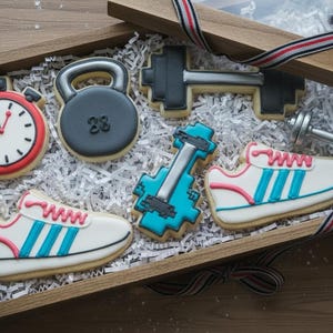 Puede incluir: Una caja de regalo de madera llena de galletas con temática de fitness. Las galletas incluyen un cronómetro, una pesa rusa con "28", una barra, una mancuerna y dos zapatillas deportivas con rayas azules y detalles rosas. La caja está forrada con papel blanco.