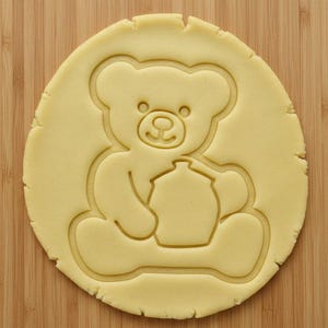 Puede incluir: Una masa de galletas redonda y de color amarillo claro con un diseño de oso de peluche presionado. El oso sostiene un tarro. La masa está sobre una superficie de madera.
