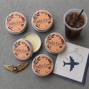 May include: An assortment of gifts for flight crew appreciation. Includes small round tins with the text "Flight Crew Appreciation", a gold-colored wing pin, a cup of soda with ice, and a napkin with an airplane illustration.