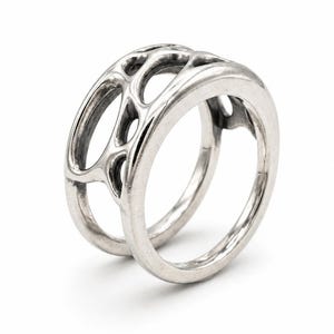 May include: A silver ring with an open, web-like design. The ring features a thick band and a series of interconnected, oval-shaped openings. The ring is presented on a white background, highlighting its intricate details and modern aesthetic.
