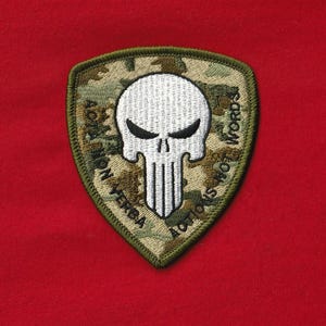 May include: Embroidered patch with a white skull design, set against a camouflage background. The patch is shield-shaped with the text "ACTIONS NOT WORDS" and "ACTA NON VERBA" in a circular pattern around the skull. The patch has a green border.