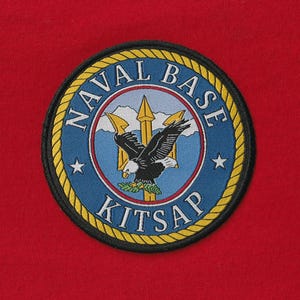 May include: Embroidered patch with a circular design featuring an eagle, trident, and the text "NAVAL BASE KITSAP." The patch has a blue and gold color scheme with a red background.
