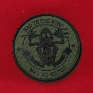 May include: A circular olive green patch with black embroidered text and a skeleton graphic. The text reads "BAD TO THE BONE" at the top and "MAL AD OSTEO" at the bottom. The skeleton is in the center, with lightning bolts on either side.