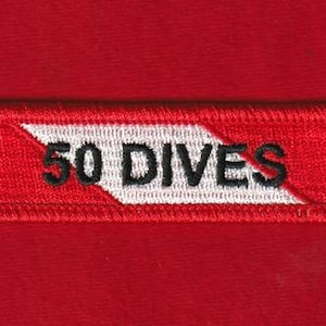 Puede incluir: Un parche rectangular rojo bordado con el texto "50 DIVES" en negro. El parche tiene una franja diagonal blanca. El parche está sobre un fondo rojo.