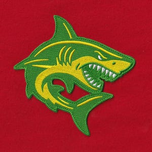 Può includere: Una toppa ricamata con il disegno di uno squalo in filo verde e giallo. Lo squalo è raffigurato con la bocca aperta che rivela denti bianchi, su uno sfondo di tessuto rosso a tinta unita. La cucitura è densa e presenta una trama visibile.
