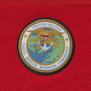 Puede incluir: Parche circular con las palabras "UNITED STATES NAVAL SPECIAL WARFARE COMMAND" que rodean un emblema central. El emblema presenta un águila, un ancla, un globo terráqueo y una figura con un tridente. El parche está sobre un fondo rojo.